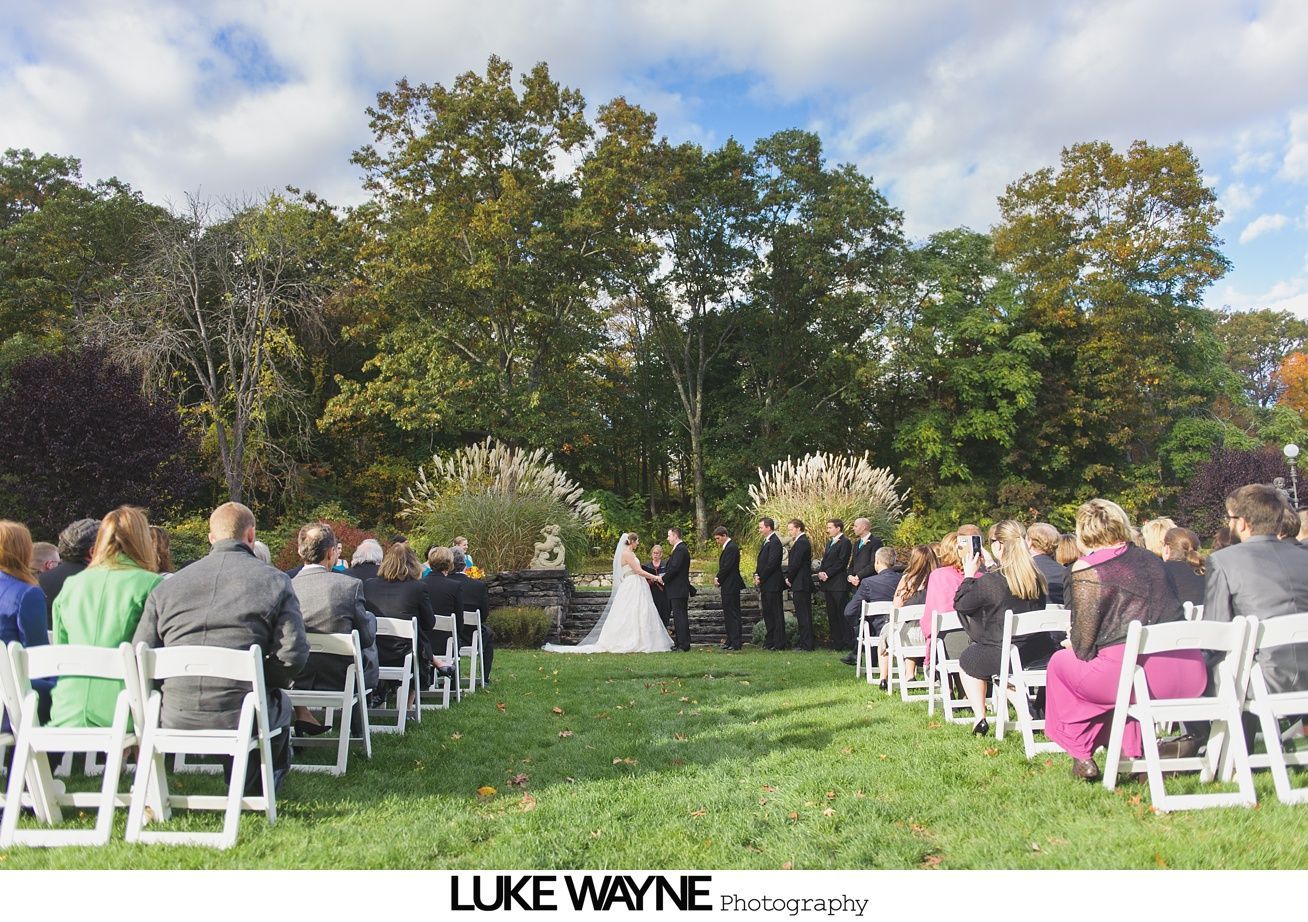 Wedding ceremony with couple, wedding party, and guests outdoors on a grassy lawn with trees and blue sky.