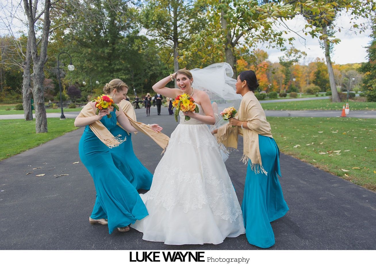 Bride and bridesmaids pose playfully on path. Bride wears white dress and veil; bridesmaids in teal dresses.