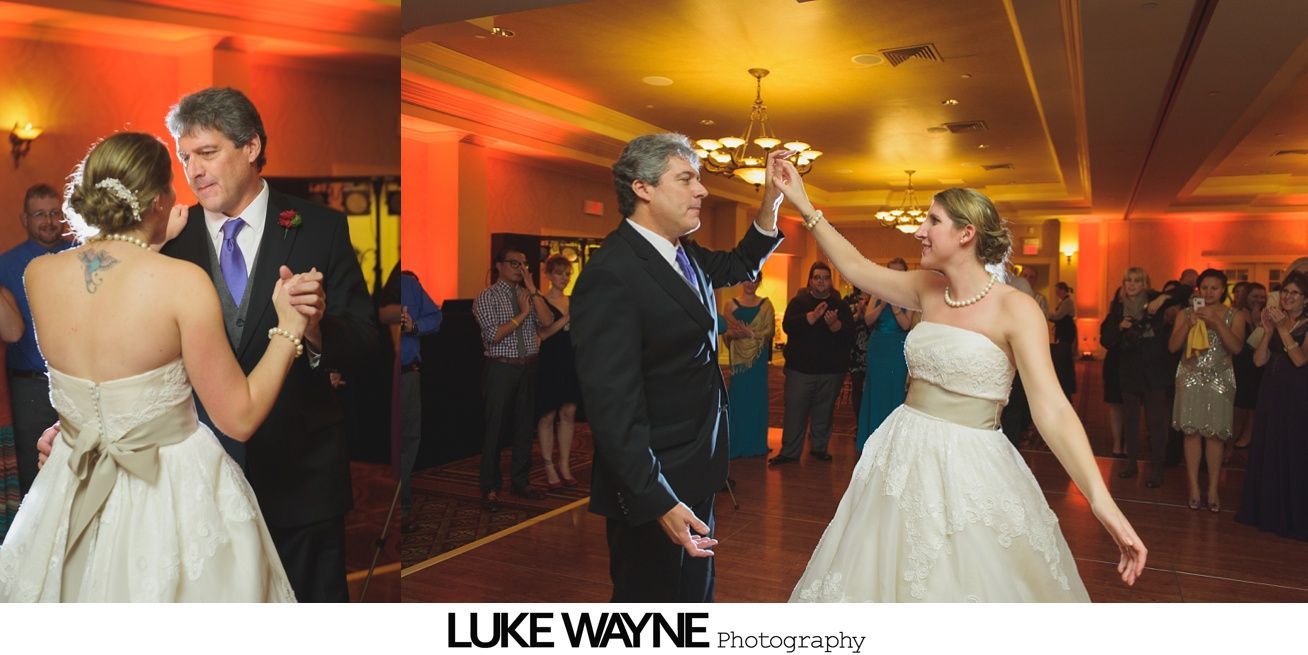 Bride and groom dance at a wedding reception with guests clapping in the background under warm lighting.