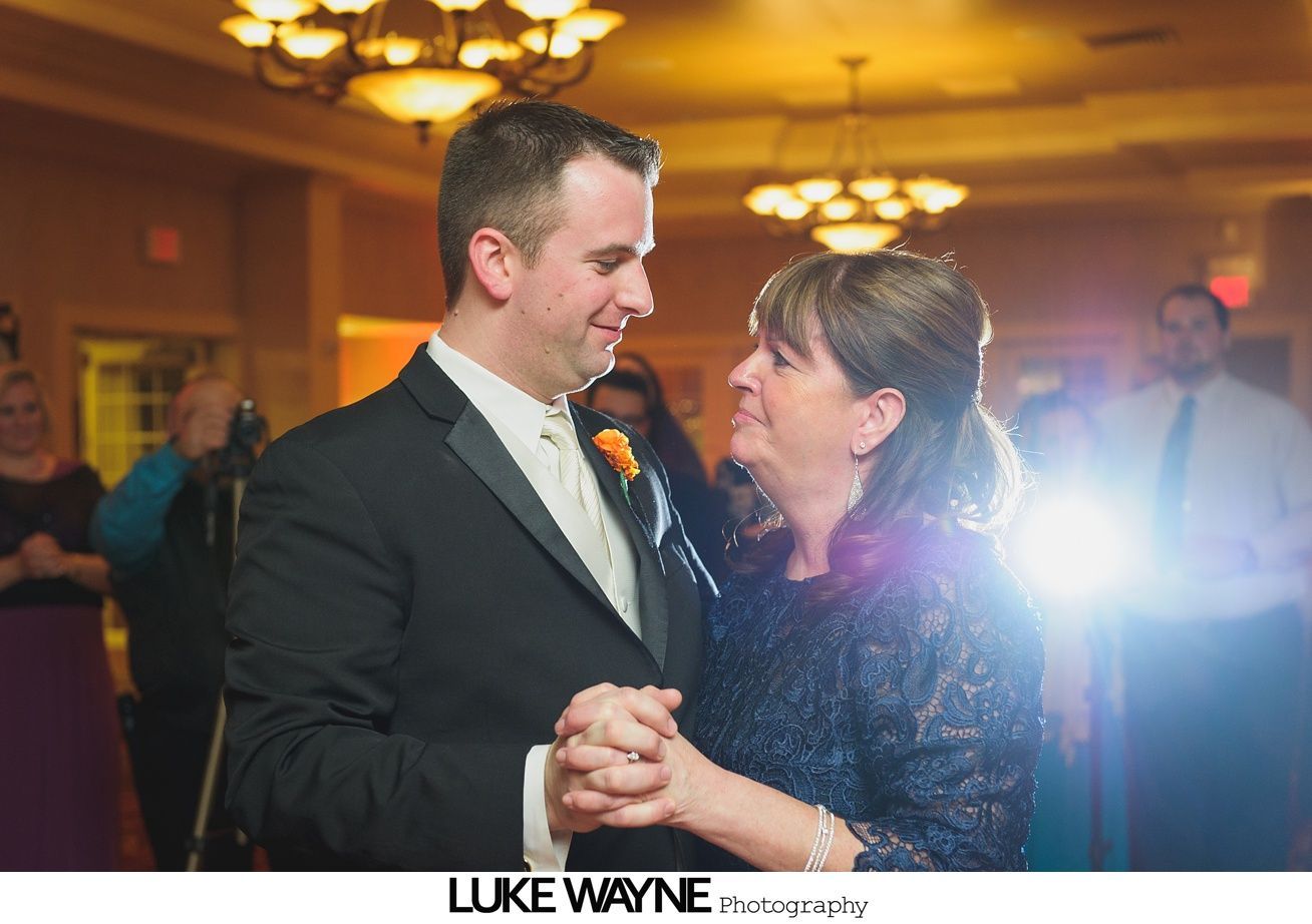 Groom and mother dancing at wedding reception, hands clasped, warm smiles, soft lighting.
