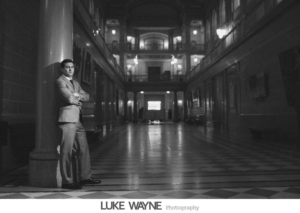 Bride in strapless white dress holding purple flowers, smiling. Dark background with archway.