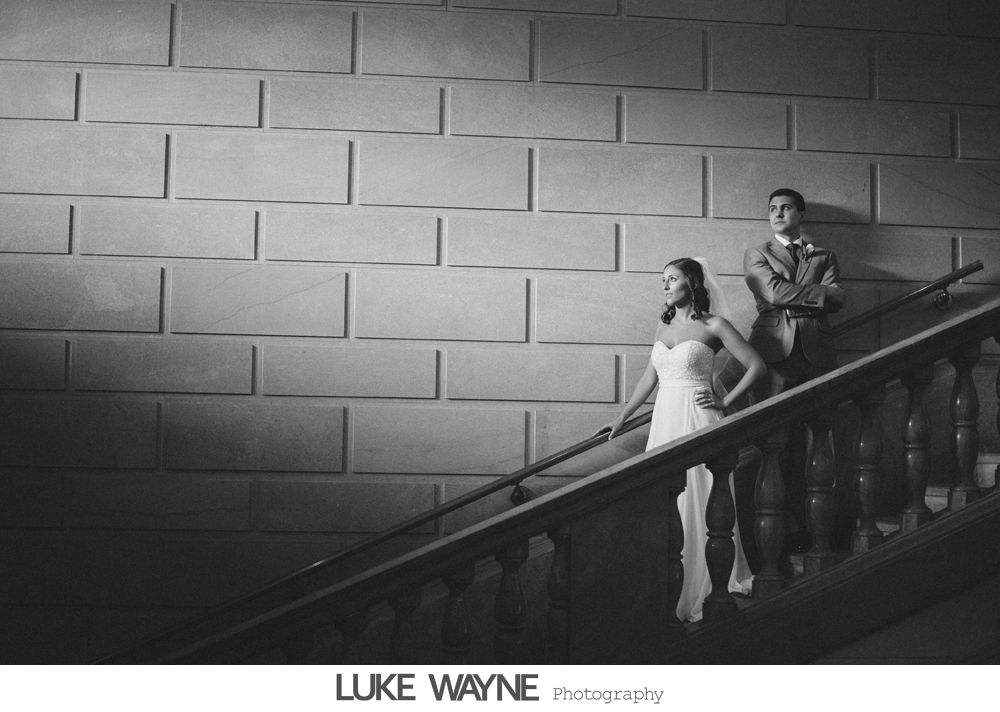 Bride and groom walk down sidewalk, groom smiles, bride holds purple bouquet, city background.
