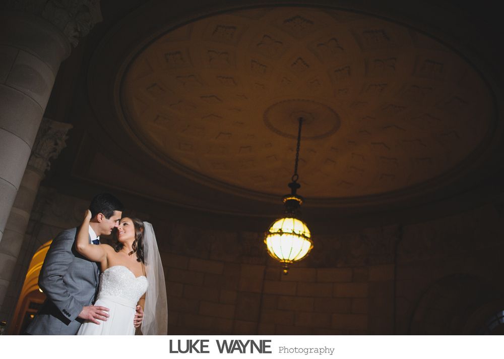 Bride and groom pose near dart boards. Bride in white dress, groom in suit. Indoor setting, red velvet ropes.