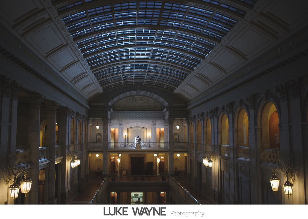 Couple in wedding attire posing in a bar. The woman's dress is white, the man's suit grey, both smiling.