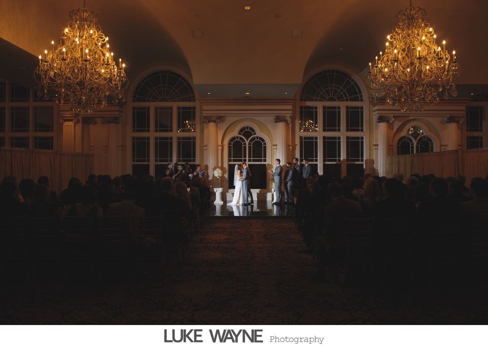 Wedding ceremony: Bride and groom exchanging rings, then kissing in front of large arched window.