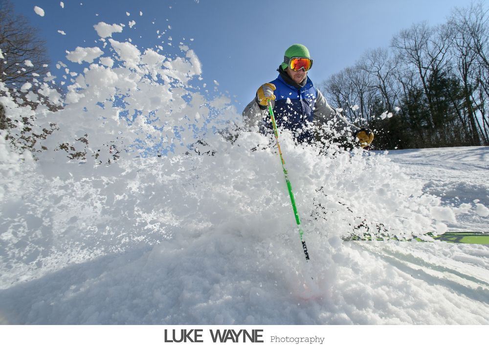 Bride in strapless white dress holding purple flowers, smiling. Dark background with archway.