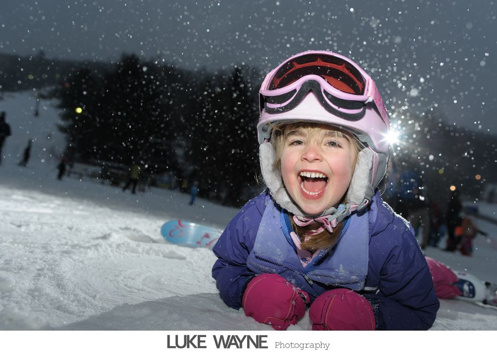 Bride in strapless white dress holding purple flowers, smiling. Dark background with archway.