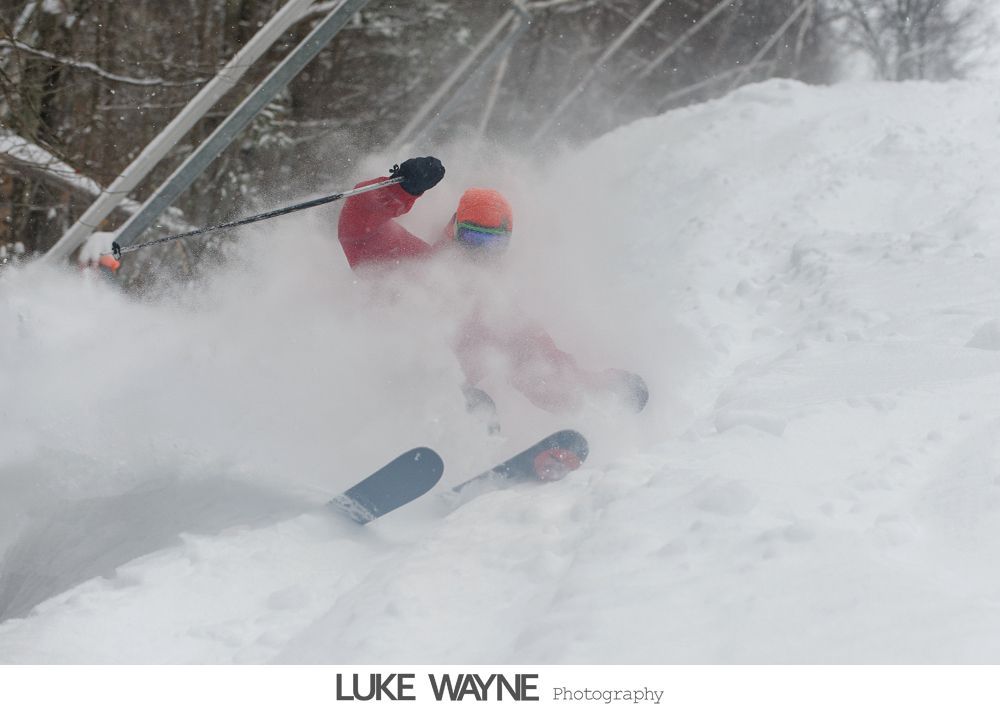 Bride and groom walk down sidewalk, groom smiles, bride holds purple bouquet, city background.