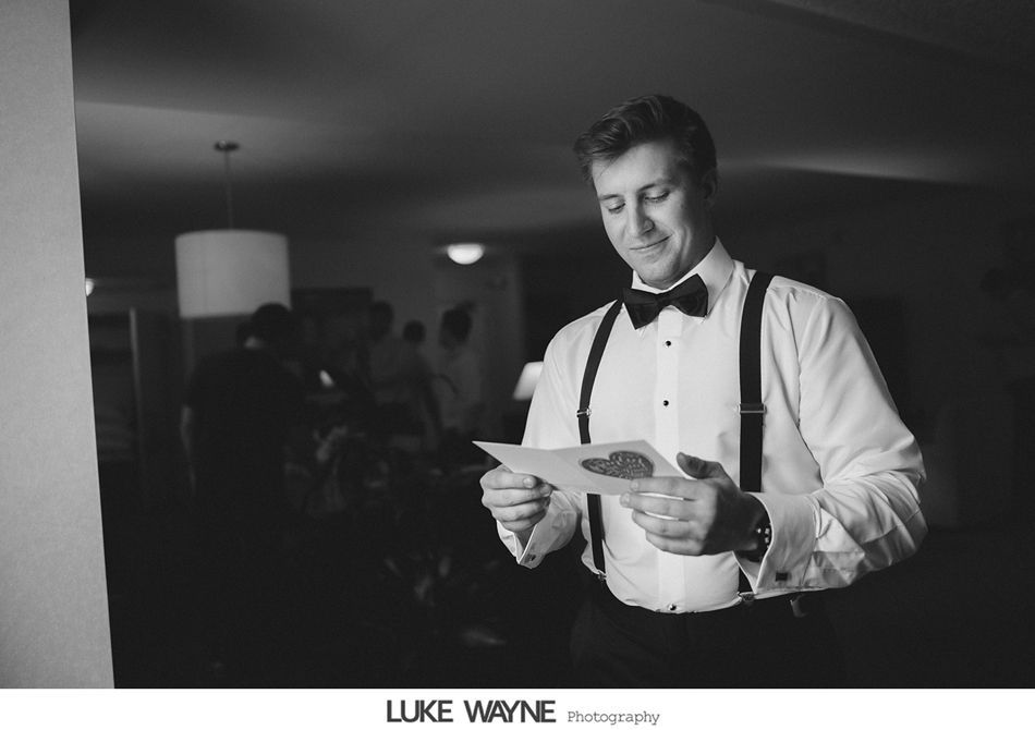 Groom reading a card, wearing a tuxedo shirt, suspenders, and bow tie, indoors. Black and white photo.