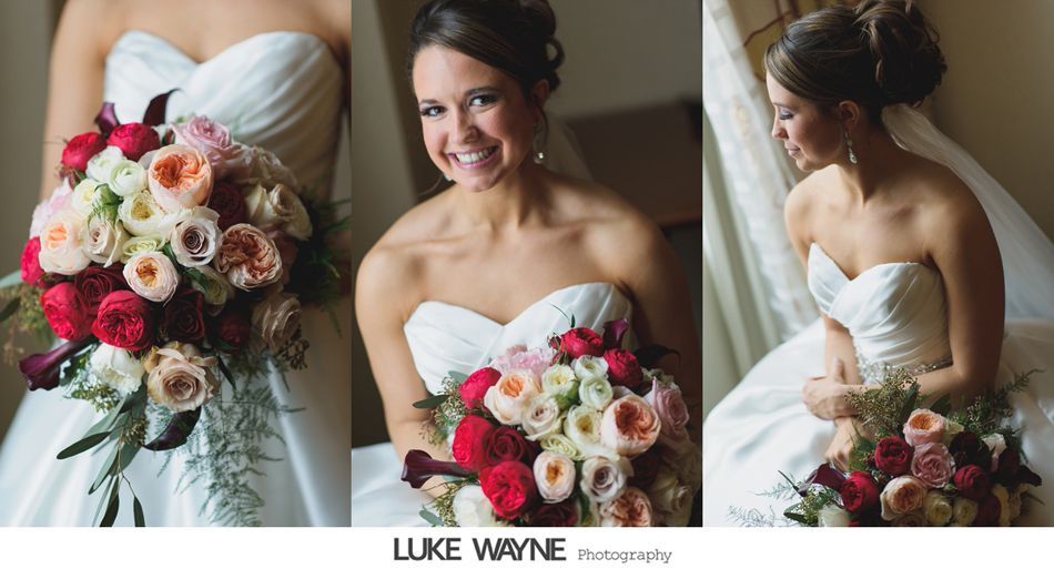 Bride in strapless gown holds bouquet of red, cream, and peach flowers, smiling in a window.