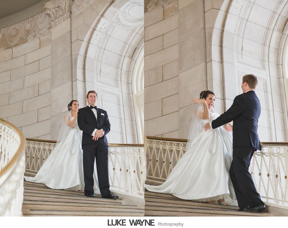 Bride and groom on a stone staircase. Groom looks on as bride adjusts her dress. White walls, gold railing.