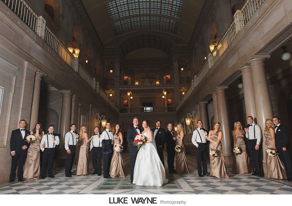 Wedding party in formal attire poses in grand hall with architectural details.