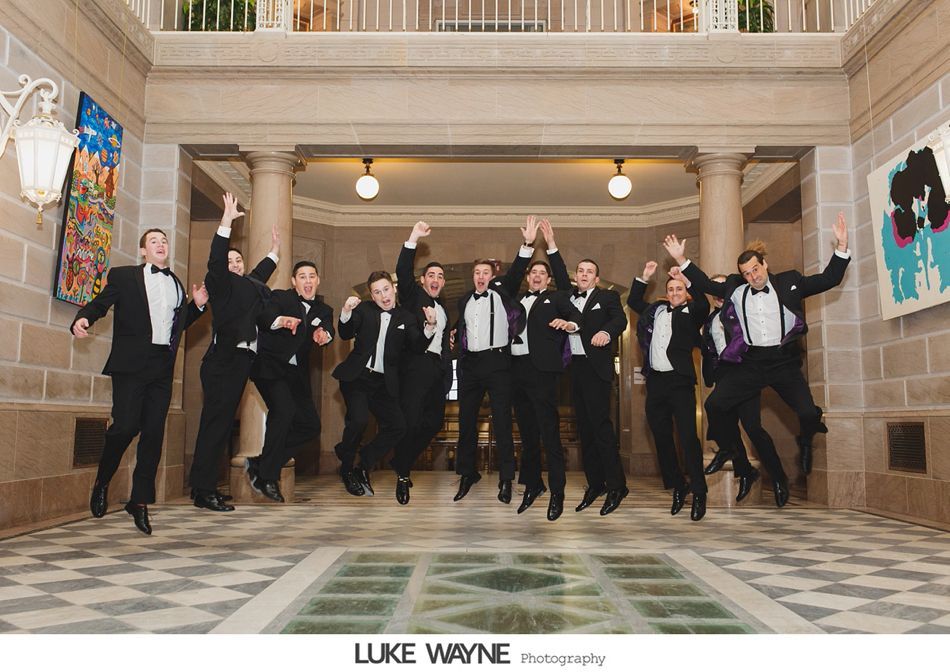 Groomsmen in tuxedos jumping for joy in a grand hall with checkered floor, and artwork.