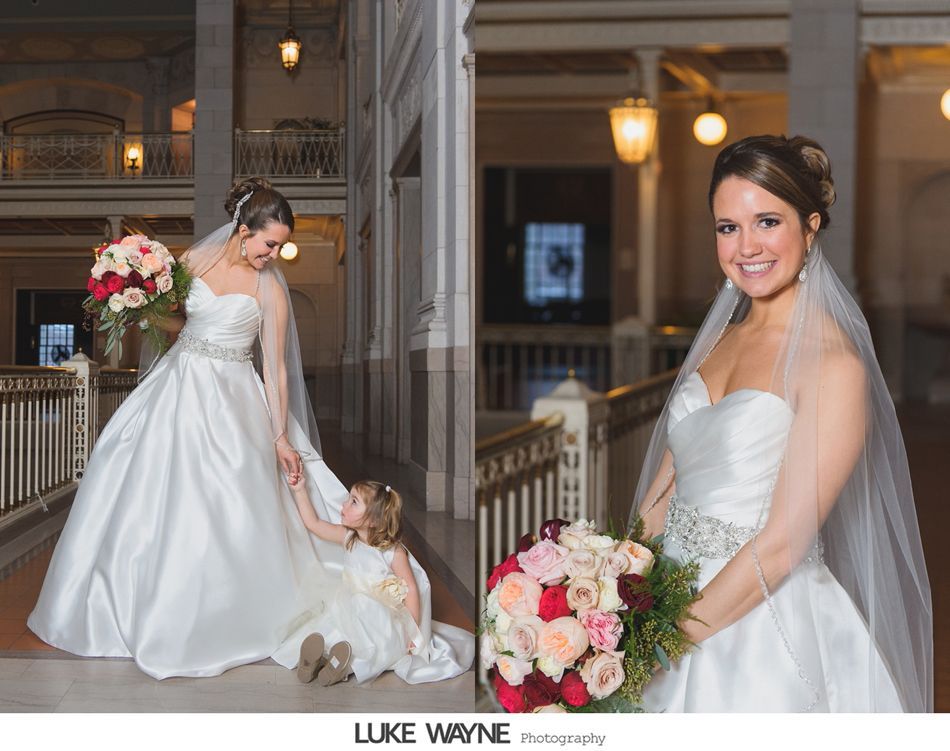 Bride in satin gown with bridesmaid, both holding bouquets, in a building with ornate architecture.