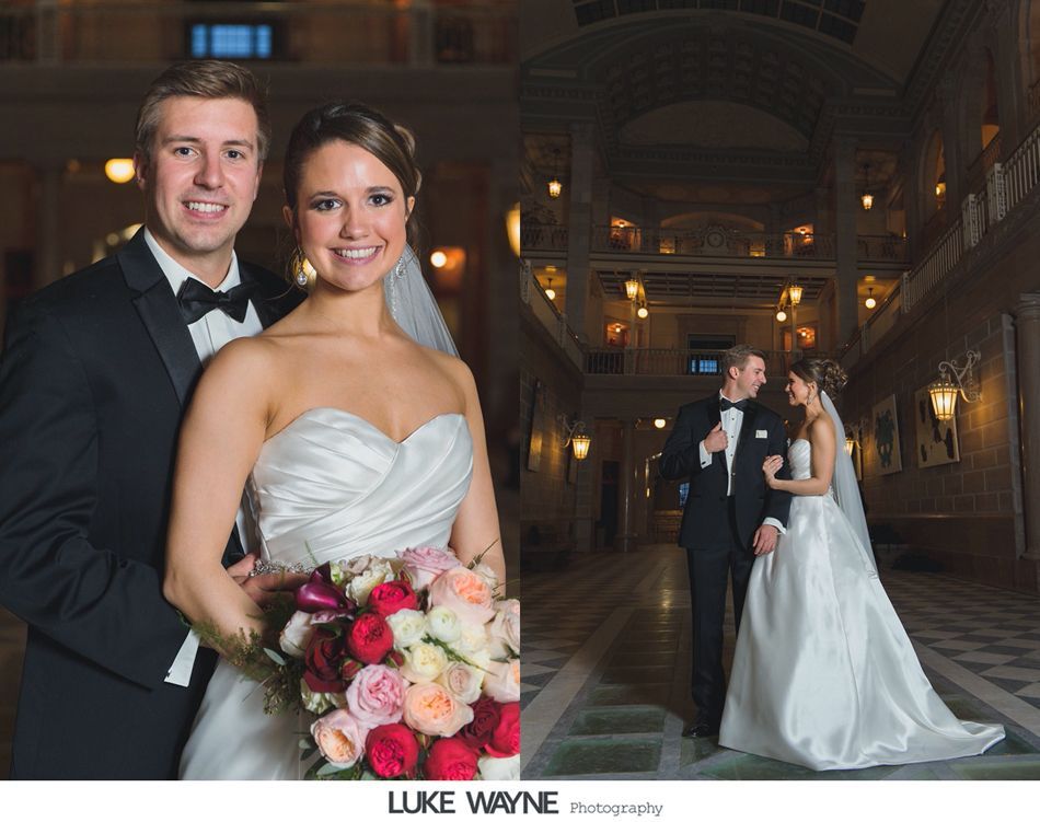 Bride and groom pose for wedding photos in a grand hall, she in a strapless dress, he in a tuxedo.