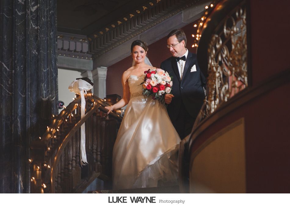 Bride and father descend a staircase, holding hands, with floral decorations and lights.