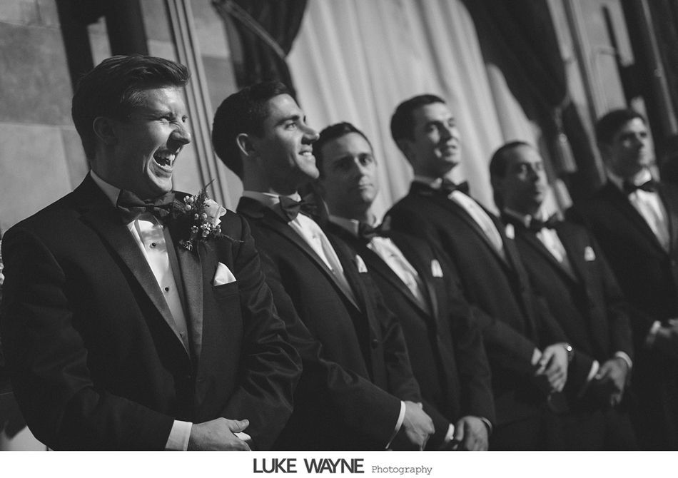 Men in tuxedos at a wedding, one with an excited expression. Black and white.