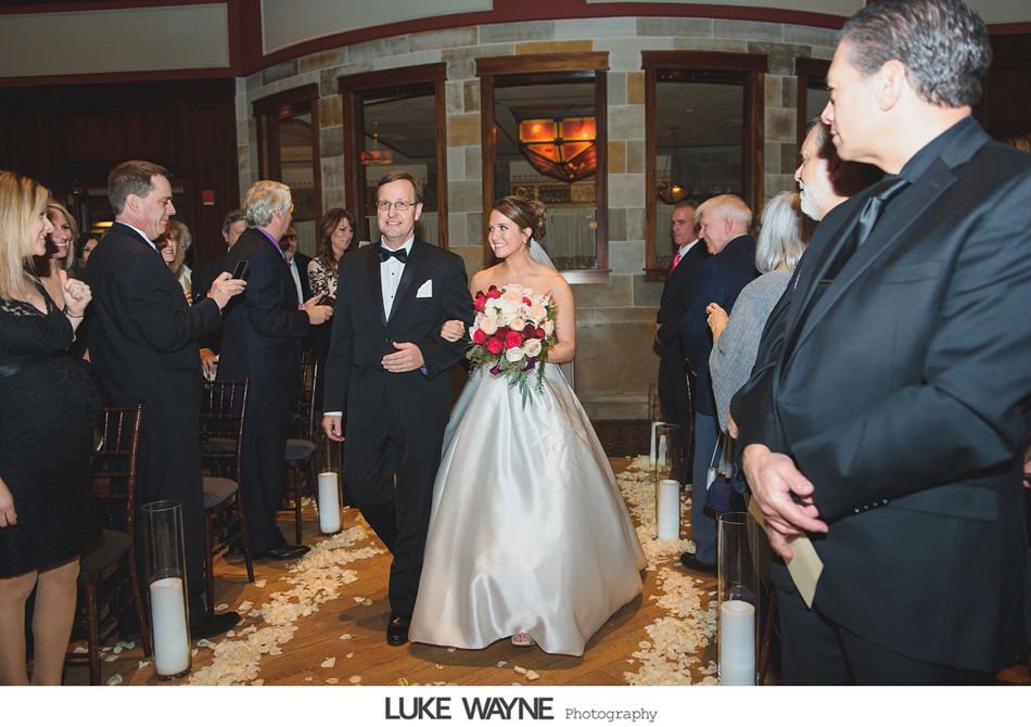 Bride and father walk down the aisle. White dress, holding flowers, surrounded by guests.