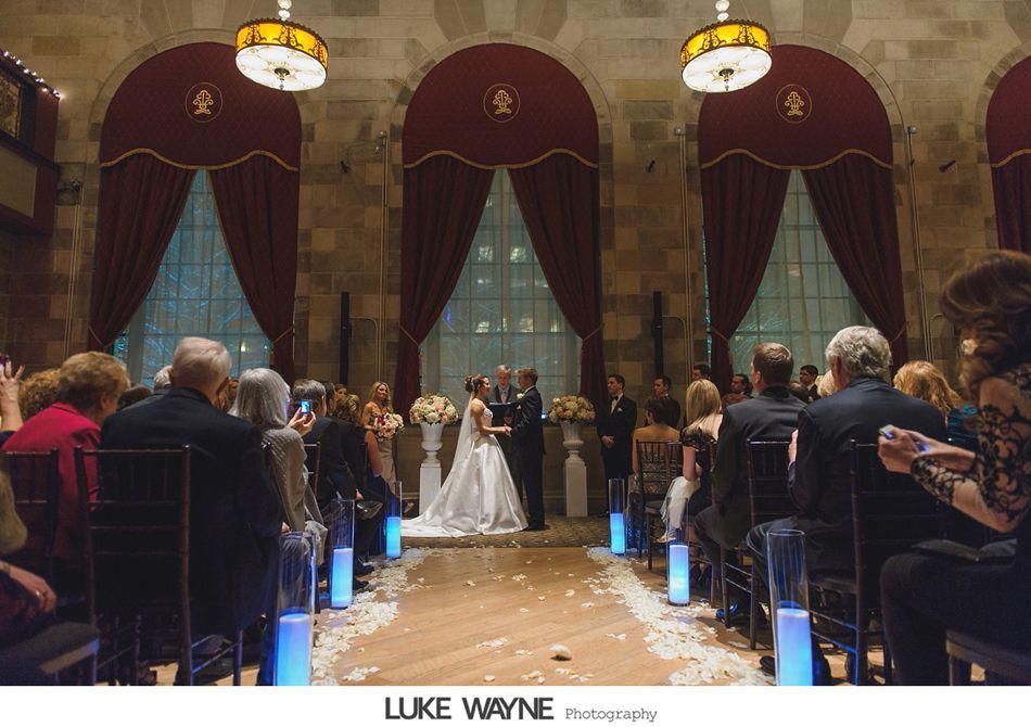 Wedding ceremony in a grand hall: couple at altar, guests seated, aisle lined with lights. Red curtains, ornate decor.