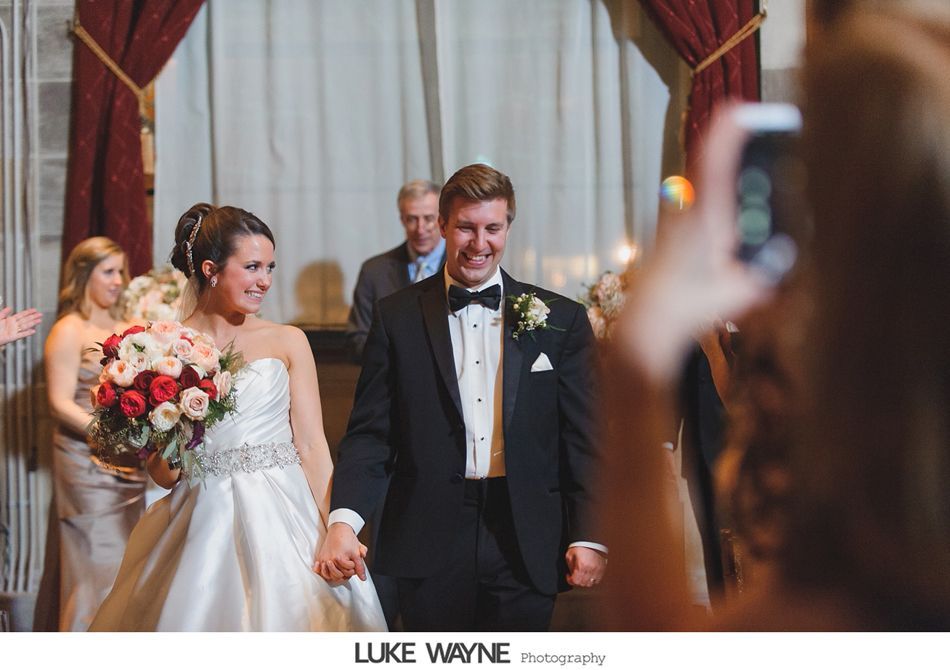 Bride and groom hold hands, smiling, exiting a ceremony. Guests take photos.