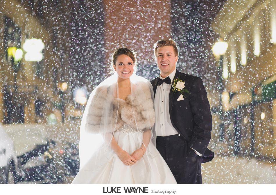 Bride and groom in formal attire, smiling in falling snow at night. City street with lights in background.