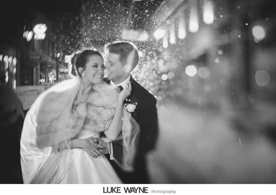 Bride and groom embrace outdoors at night; snow falls, soft lights glow.
