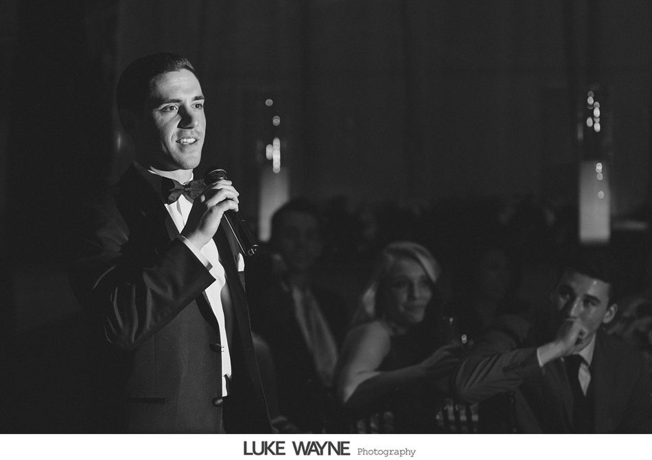 Man in suit and bow tie speaks into microphone at a formal event, with blurred people in the background.