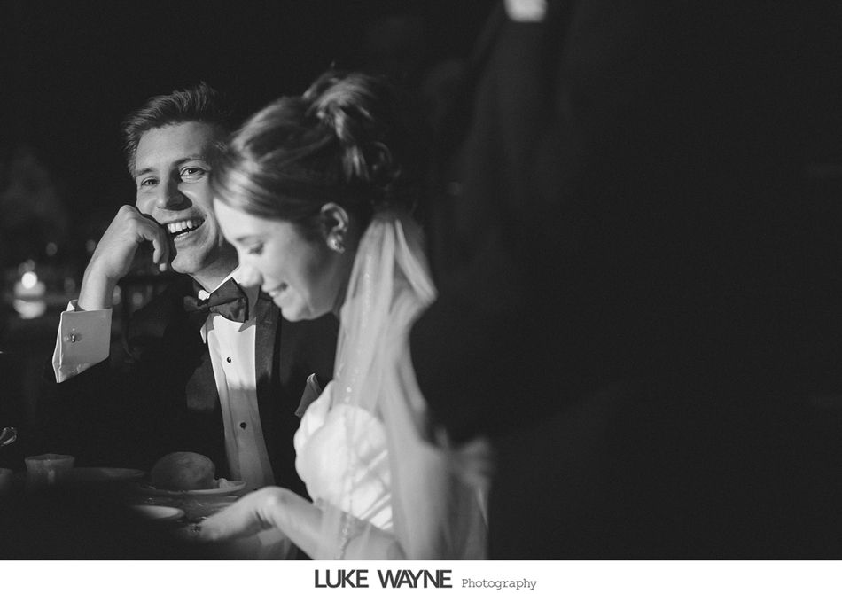 Groom laughs, bride smiles; both sit at a table, likely a wedding reception. Black and white photo.