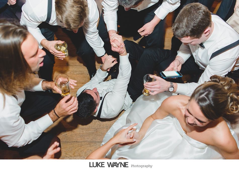 Groom lies on dance floor, surrounded by groomsmen, bride laughs, all holding drinks, wedding reception.