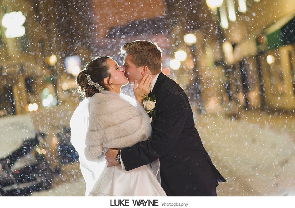 Couple kissing in the snow outside after their wedding. Bride wears a fur shawl, groom in a tuxedo, city lights in background.