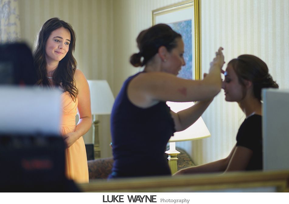 Woman in peach dress watches as stylist applies makeup to another woman in a black top. Hotel room.
