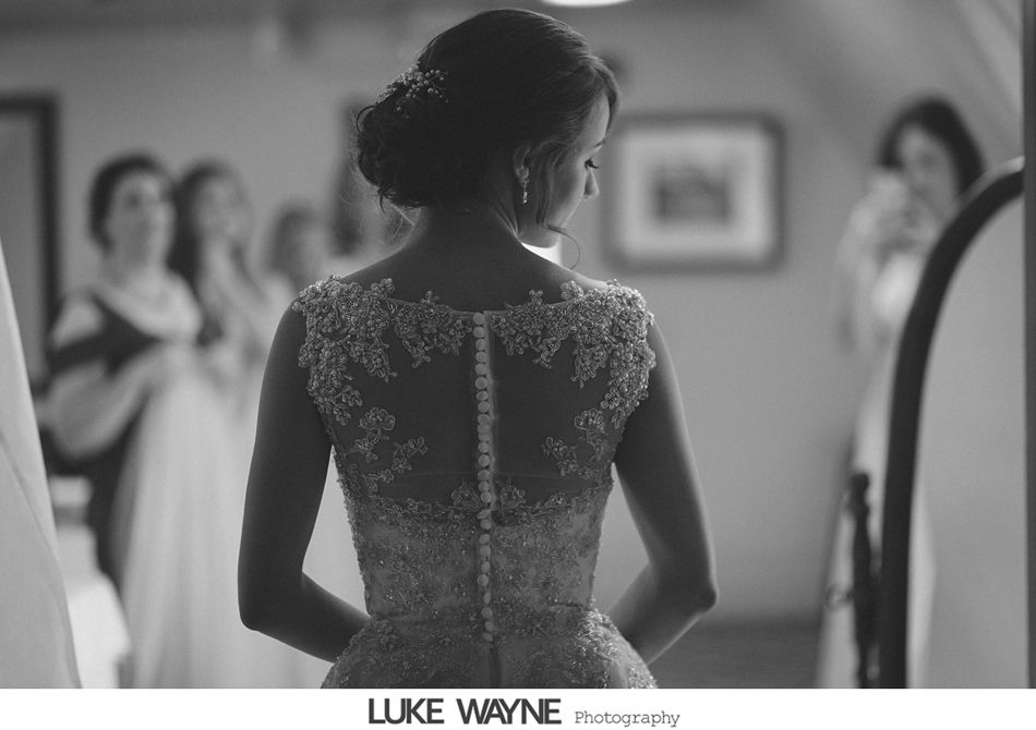 Bride in wedding dress, back to camera, surrounded by bridesmaids, looking into mirror.
