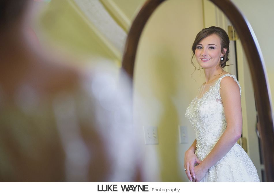 Bride in a white wedding dress smiles at her reflection in a framed mirror.