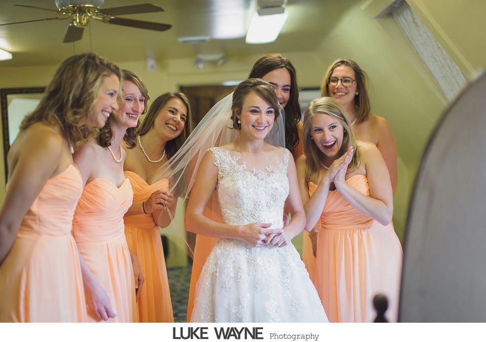 Bride in wedding dress surrounded by bridesmaids in peach dresses; looking into a mirror.
