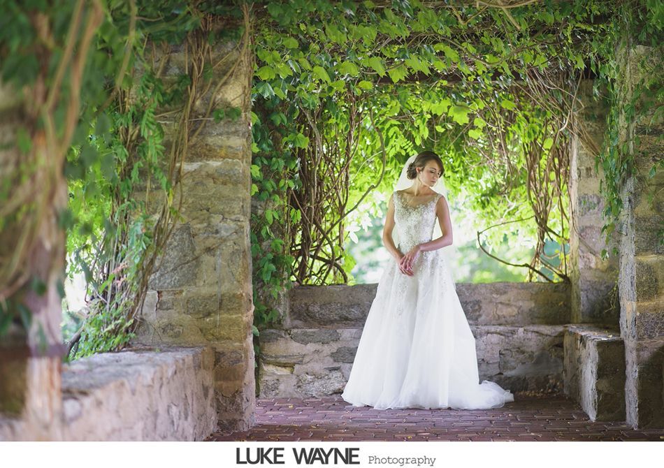 Bride in white dress under a stone and vine covered archway, looking down, smiling.