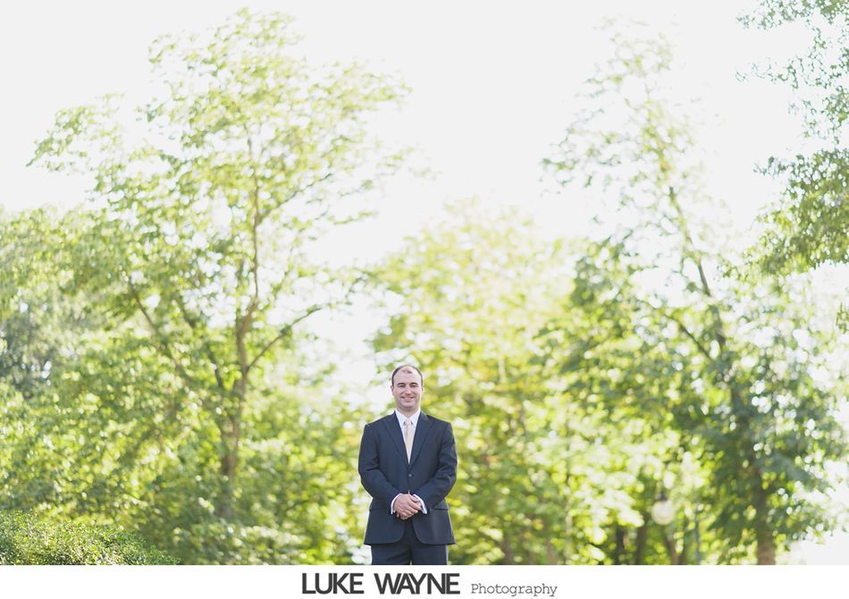 Man in suit stands outdoors, arms crossed, smiling, sunlight through trees.