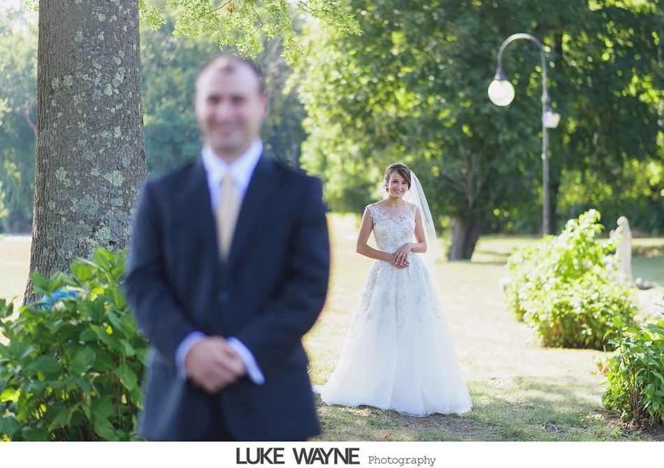 Groom blurred in the foreground, bride in white dress smiling in focus, outdoor wedding setting.