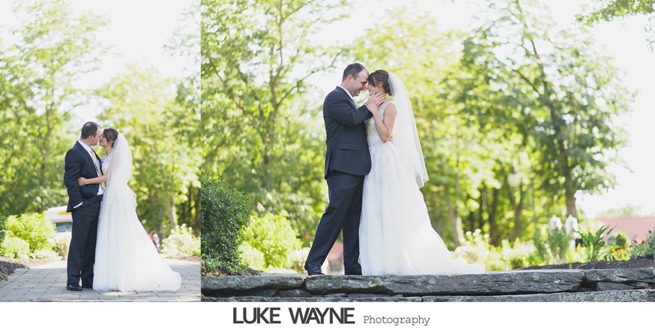 A bride and groom embrace outdoors, near lush greenery. The groom is in a suit, and the bride in a flowing white dress and veil.