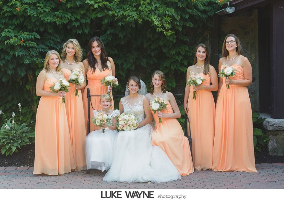 Bridesmaids in peach gowns with bouquets pose with the bride and flower girl in front of a green bush.