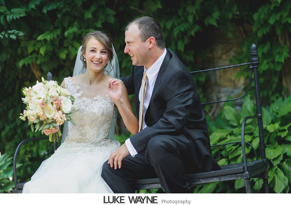 Bride and groom seated, laughing on bench, floral bouquet. Green foliage background.