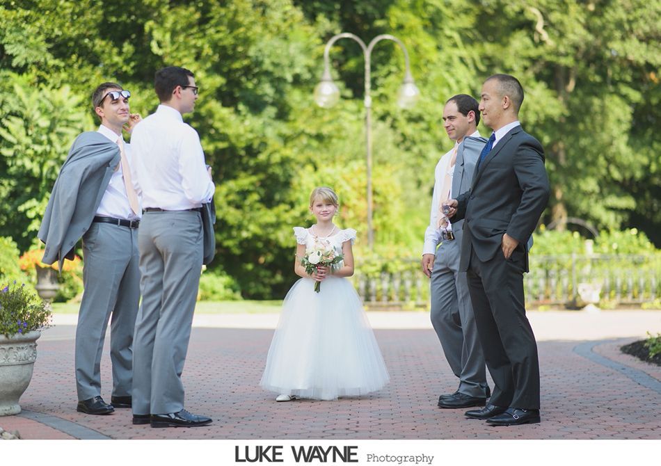 Groomsmen in gray suits stand around flower girl in white dress on a brick path, outdoors.