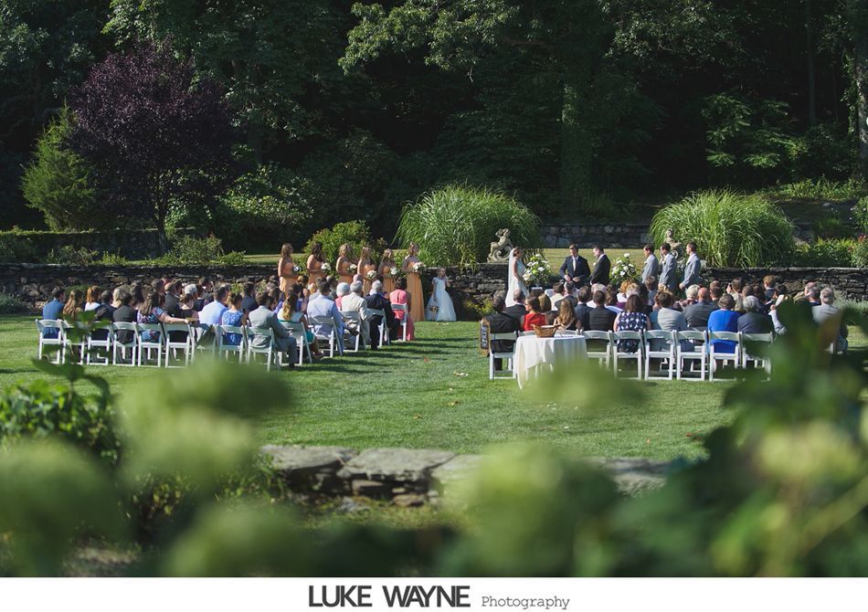 Wedding ceremony in a garden setting; guests seated, wedding party and couple stand under trees.