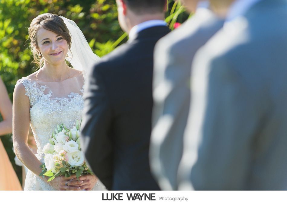 Bride smiling, holding bouquet, looking at groom during outdoor wedding ceremony.