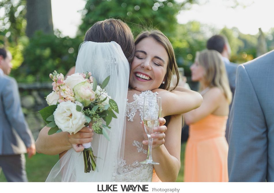 Bride hugs a smiling woman; both are outdoors. Bride holds bouquet and champagne glass. Other guests in background.