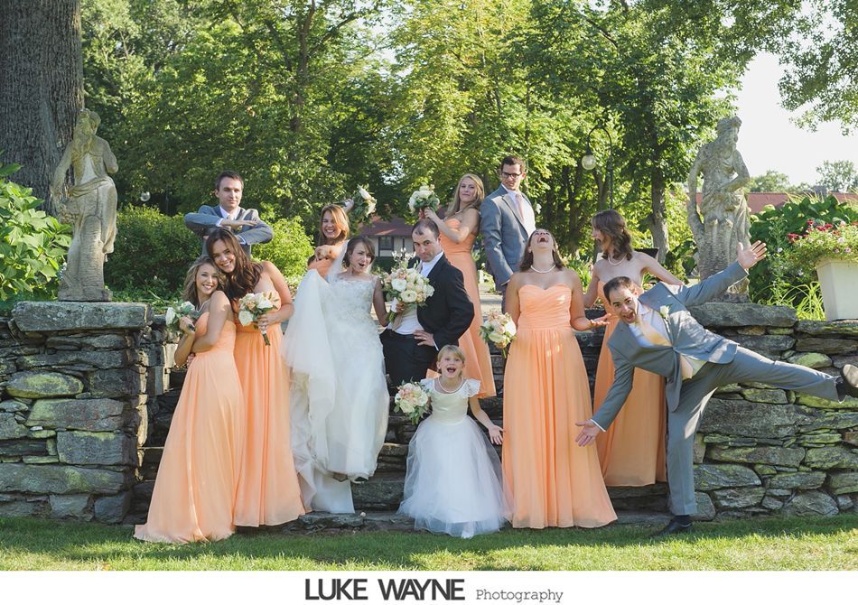 Wedding party on stone steps, bride and groom centered with bridesmaids in peach dresses, sunny outdoor setting.