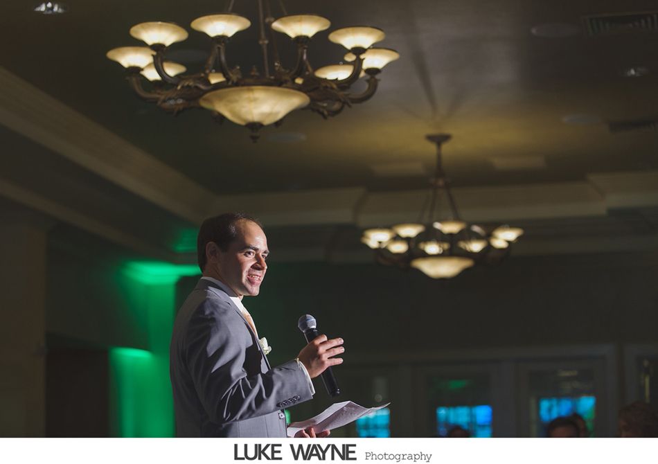 Man in grey suit speaking into microphone, holding papers under ornate chandeliers. Green background.