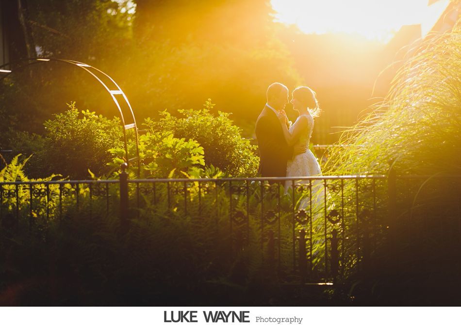 Bride and groom embrace in golden sunlight by a garden fence and arched structure.