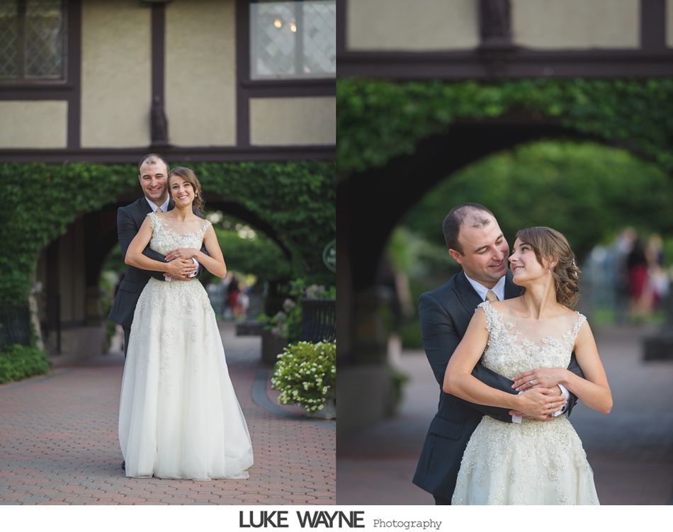 Bride and groom embrace beneath a green archway. She smiles, he looks at her, both in formal attire.
