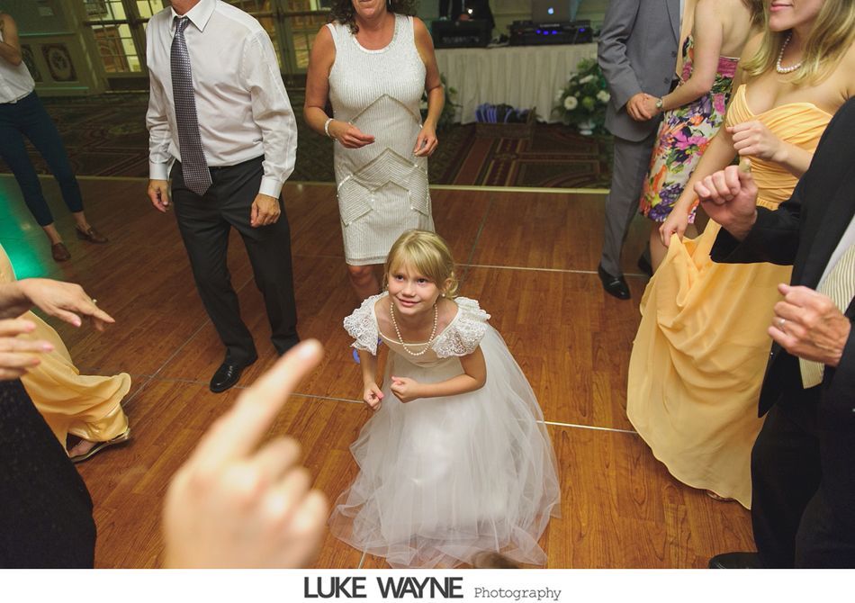 Young girl in a white dress dancing on a wooden dance floor, surrounded by other people at a formal event.