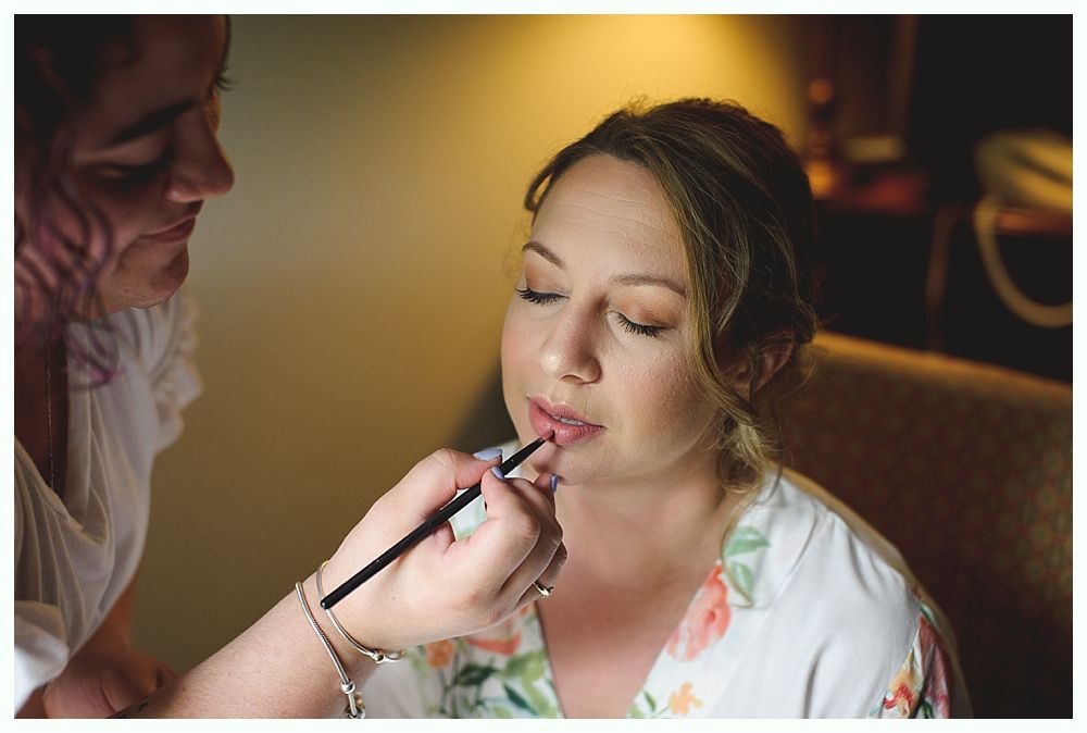 Woman having makeup applied with lip brush in warm-lit room, wearing floral robe.